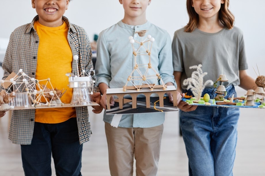 Three smiling children holding art projects made out of wooden dowels