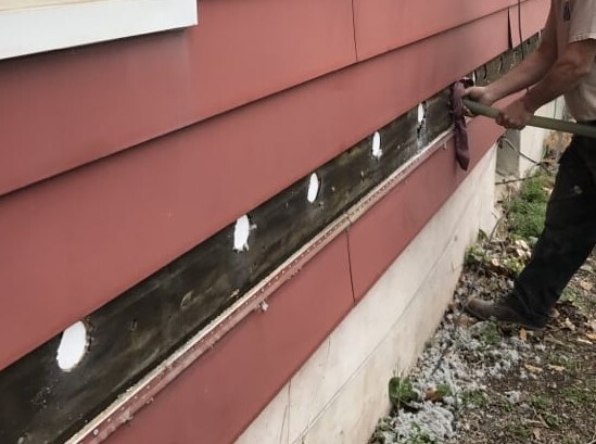 A man holding a hose to a hole on the side of a house as it injects insulation into the wall