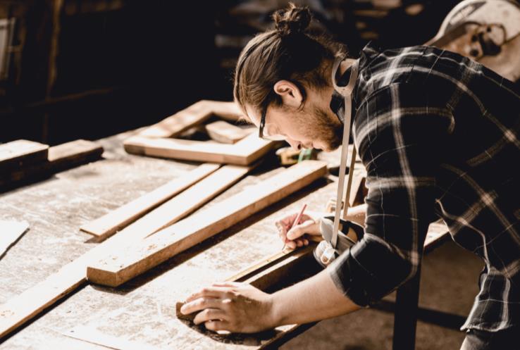 a man wearing safety goggles measures and marks a board on a work bench